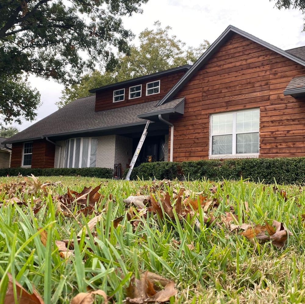 House with wooden exterior, ladder placed against the roof, and fallen leaves in the grass.