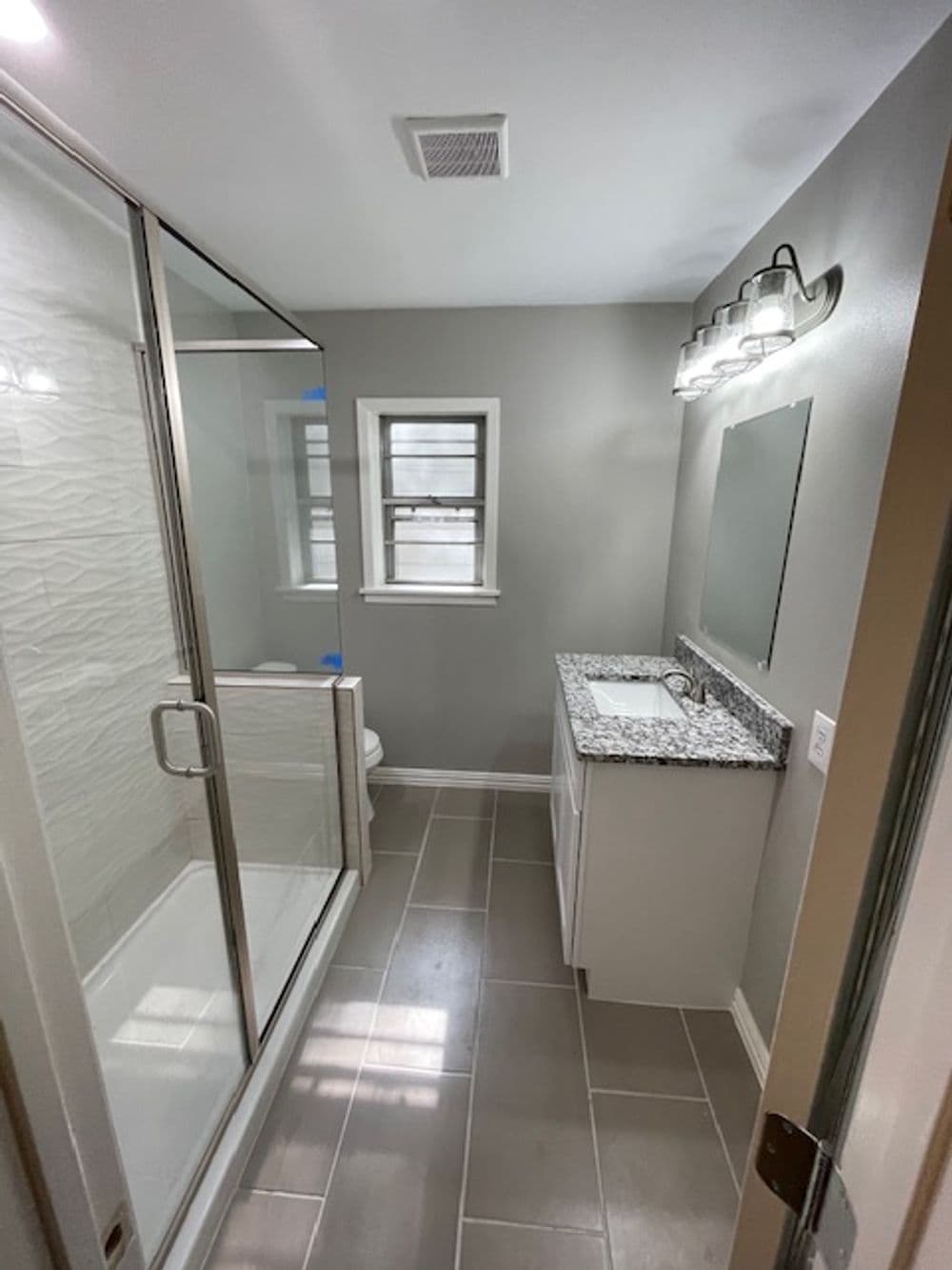 Modern bathroom with glass shower, granite countertop, and gray tile flooring. Bright lighting.