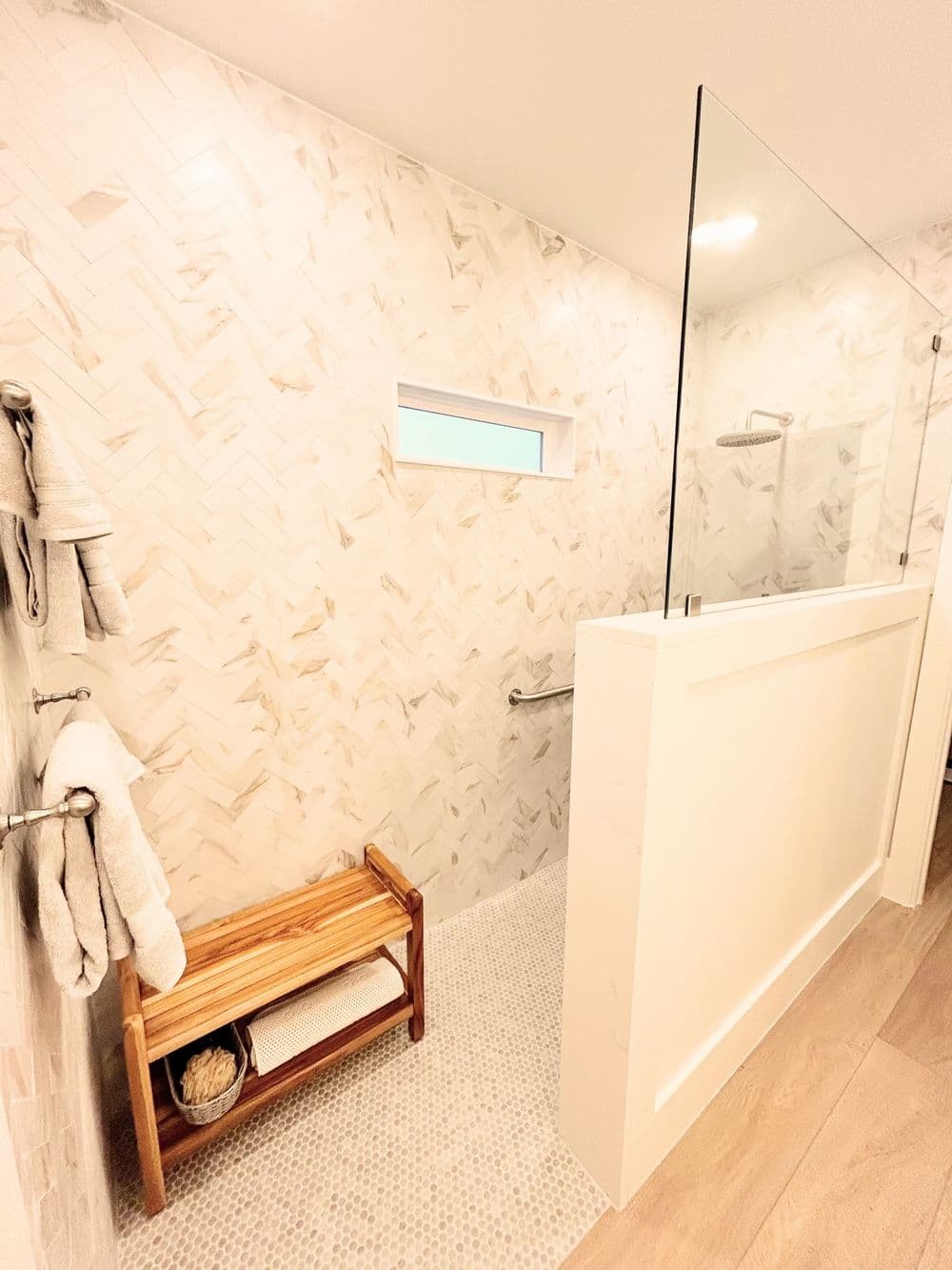 Modern bathroom shower with herringbone tiles, glass partition, and wooden bench.