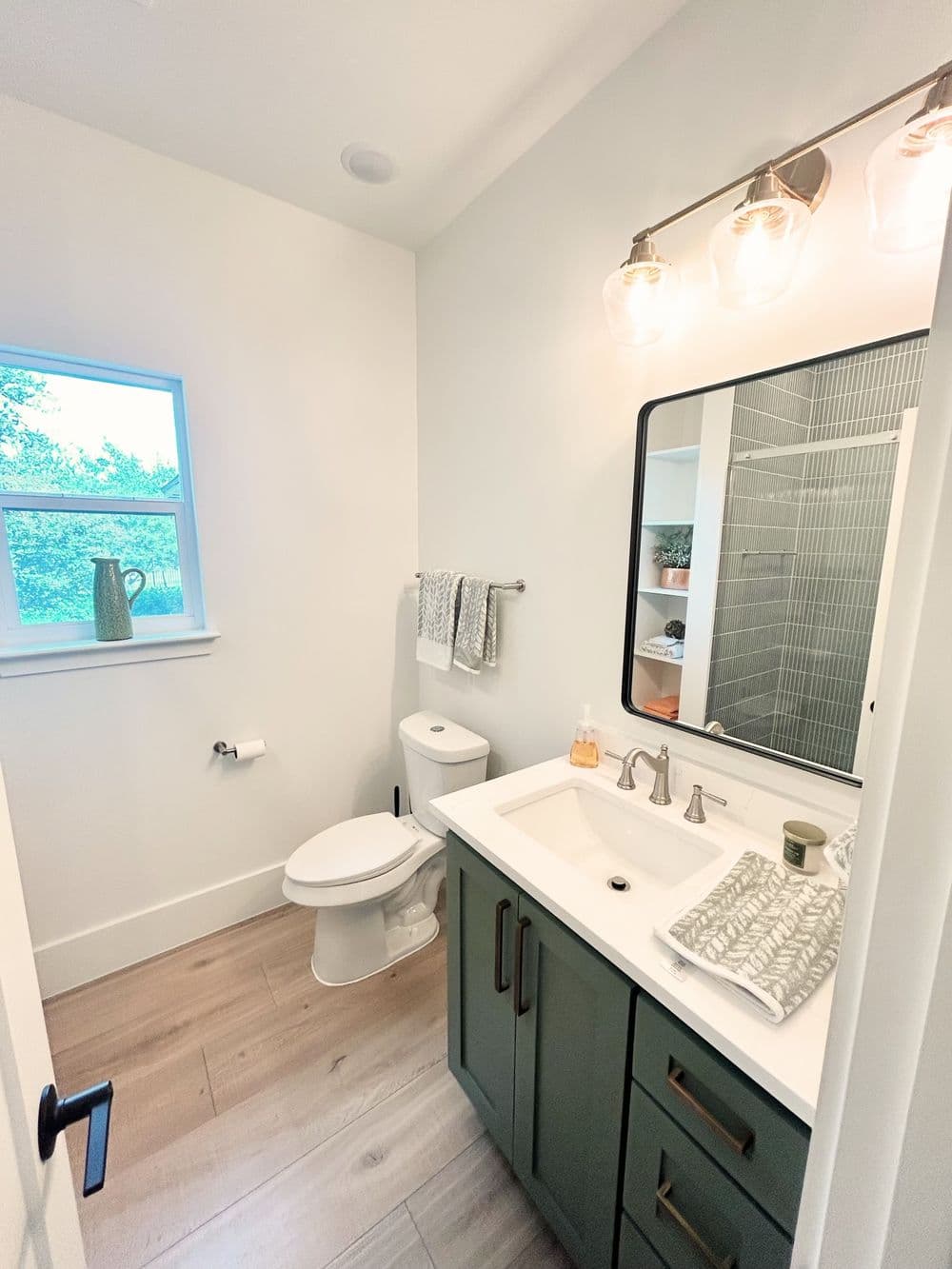 Modern bathroom with green cabinetry, large mirror, and neutral decor, featuring natural light.