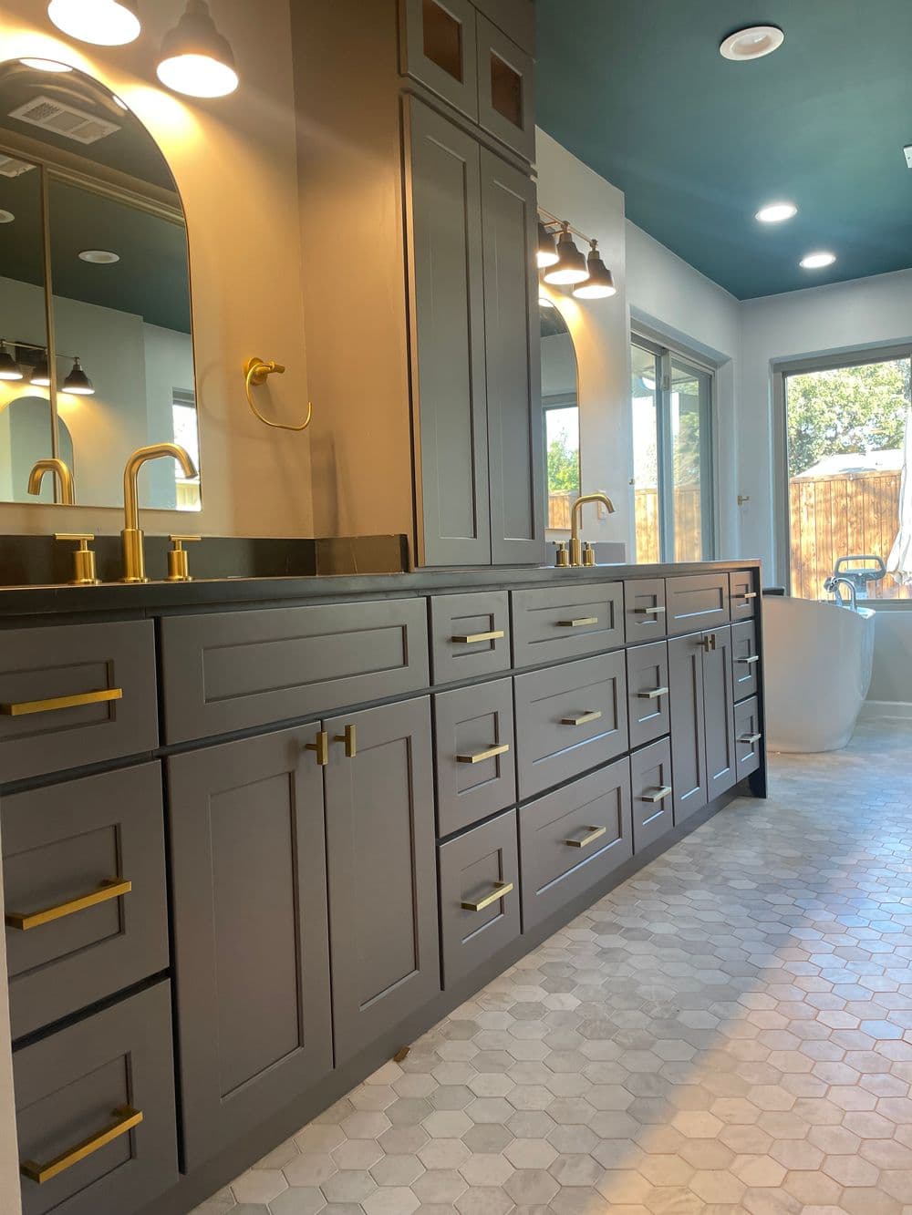 Modern bathroom with gray cabinetry, gold fixtures, and a freestanding bathtub. Natural light.