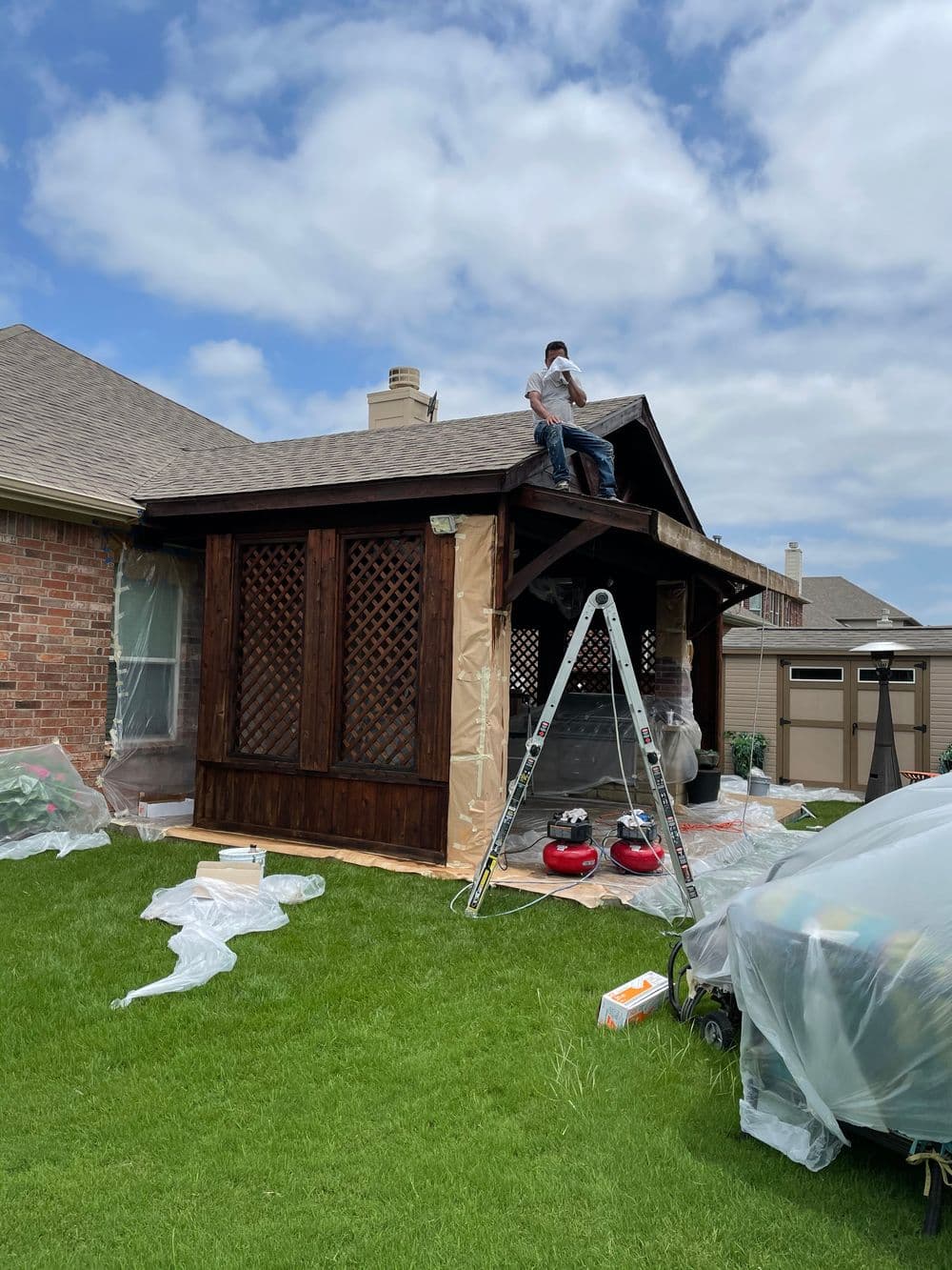 Construction worker on roof of wooden porch with lattice, surrounded by tools and protective coverings.