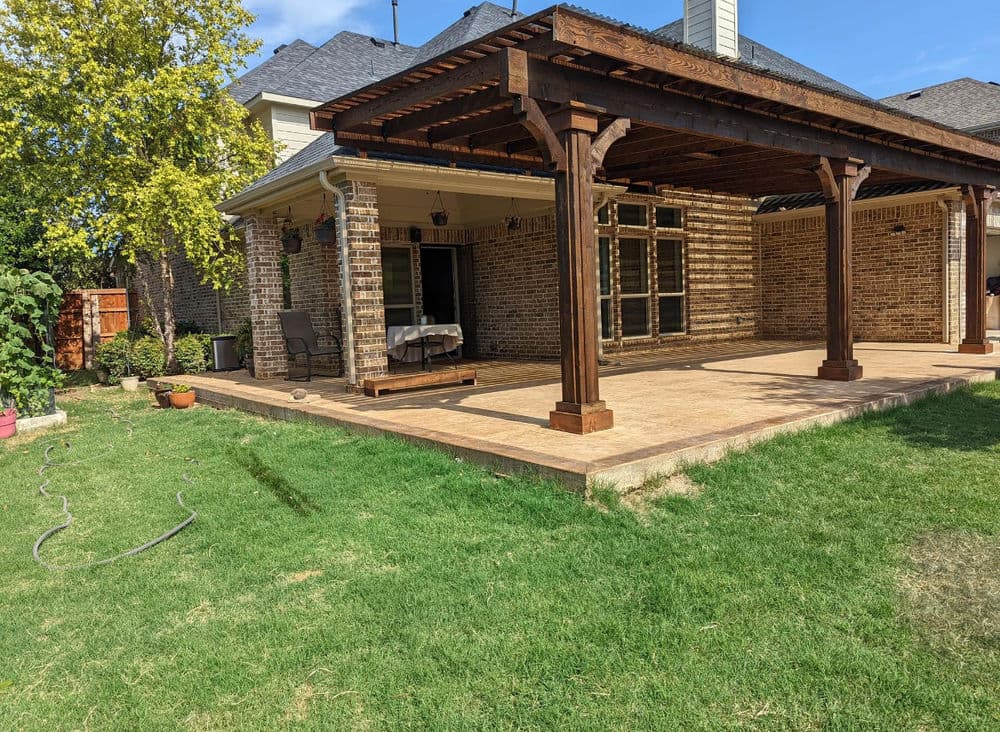 Backyard patio with wooden pergola, stone pavers, and green lawn under blue sky.